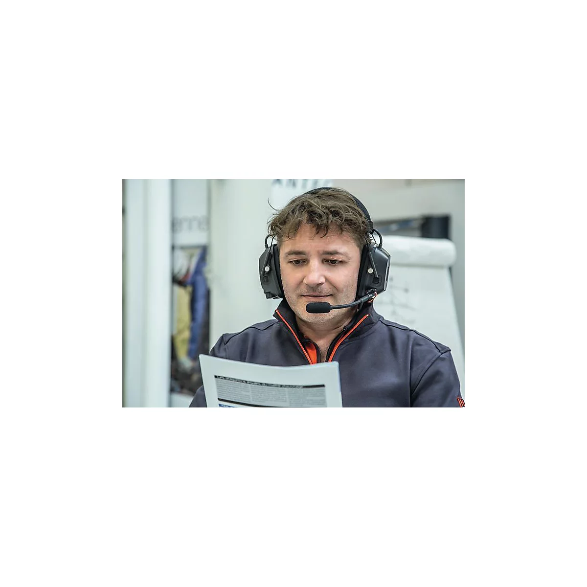 Un homme portant un casque lit un document, les yeux baissés. Il porte un sweat-shirt bleu foncé avec un accent orange.