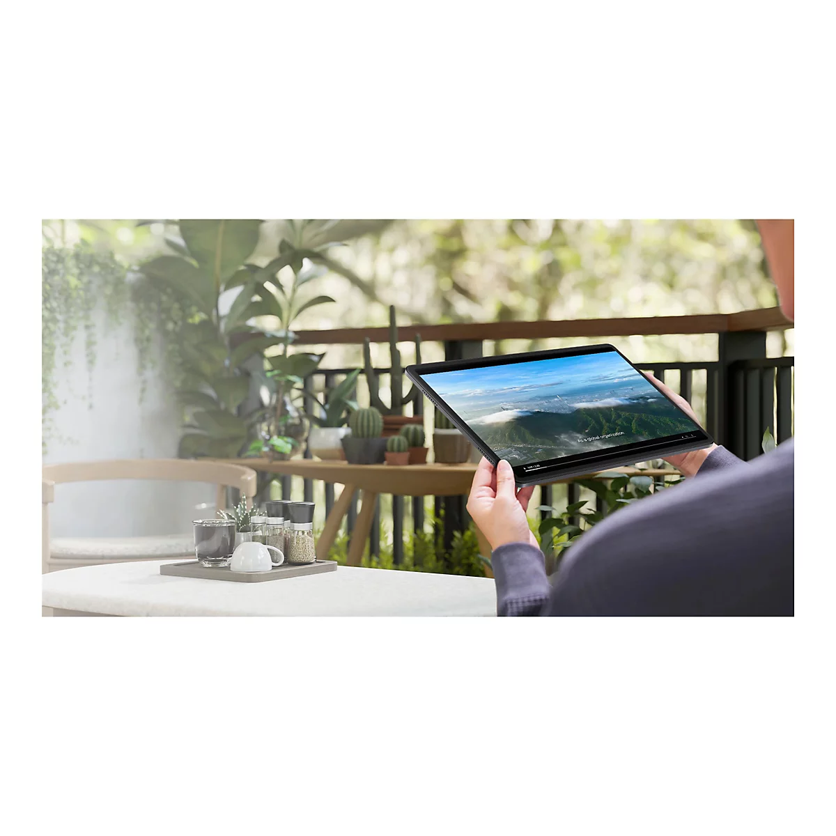 Person hält Tablet, das ein Video mit Landschaftsaufnahmen zeigt. Auf einem Tisch ist ein Kaffee-Set platziert.