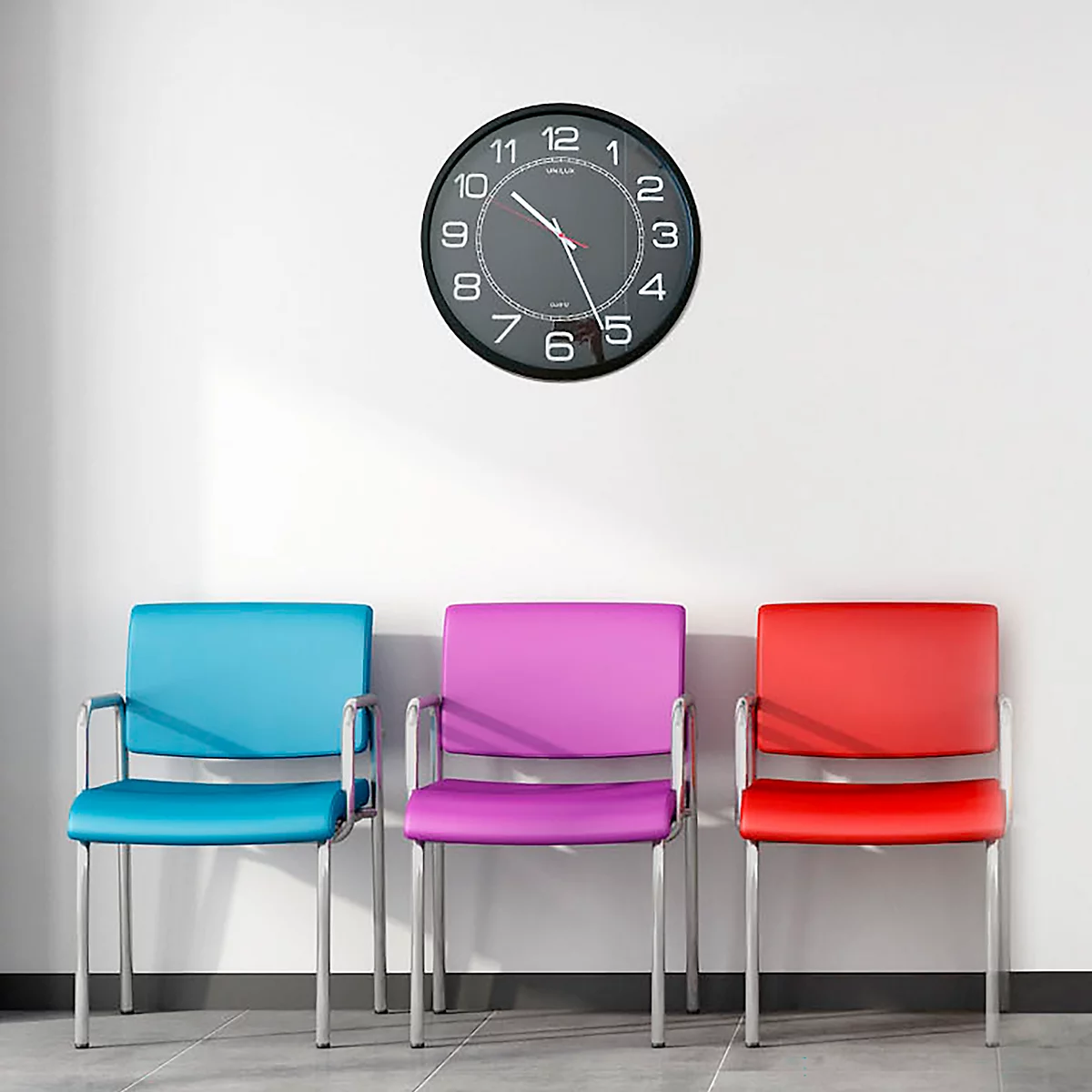 Trois chaises colorées devant un mur blanc, au-dessus d'une horloge.
