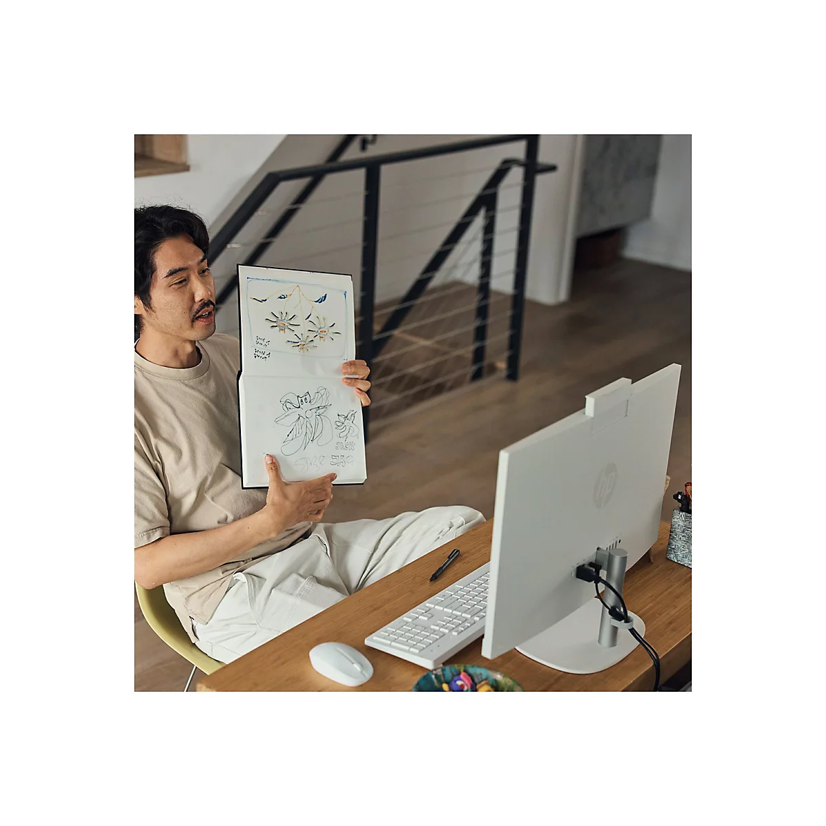 Ein Mann zeigt ein Skizzenbuch vor einem HP Computer. Er sitzt am Tisch. Auf dem Tisch sind Tastatur, Maus und Stift.