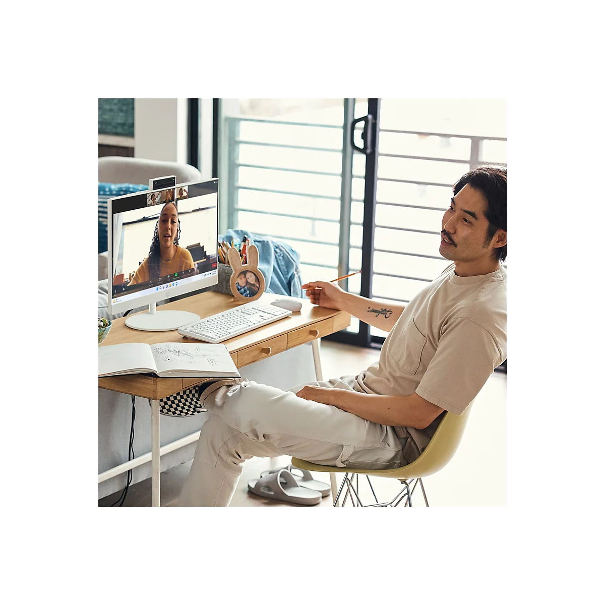 Ein Mann sitzt an einem Schreibtisch, nimmt an einem Videogespräch teil. Auf dem Monitor ist eine Frau zu sehen, die spricht. Er hält einen Stift, vor ihm liegt ein Notizbuch.