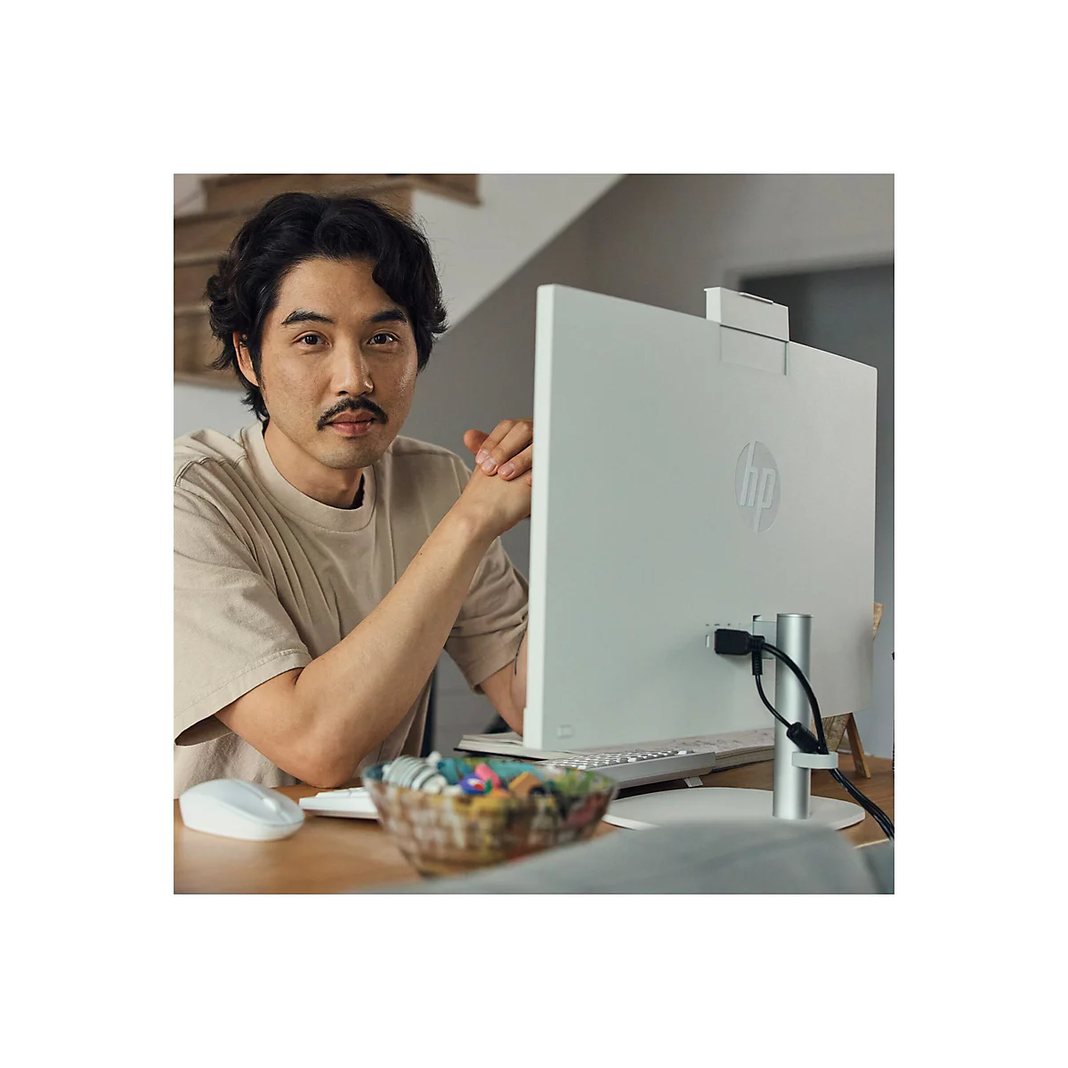 Ein Mann sitzt an einem Tisch vor einem weißen HP-Monitor. Er hat einen Schnurrbart und einen Blick in die Kamera.