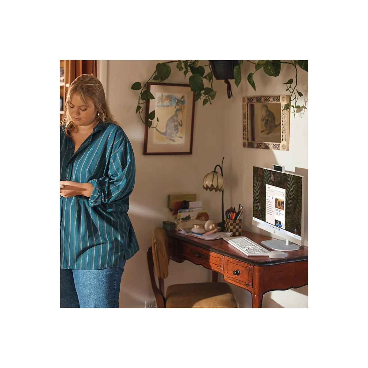 Eine Frau steht in einem Zimmer neben einem Schreibtisch. Sie trägt ein blaues gestreiftes Hemd und Jeans. Auf dem Schreibtisch steht ein All-in-One-Computer.