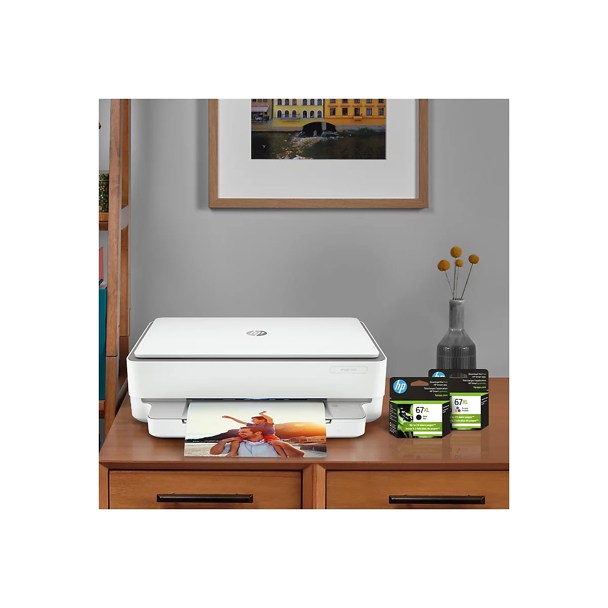 Ein weißer HP Envy 6055-Drucker steht auf einem braunen Holztisch, darunter ein Farbfoto. Rechts stehen Tintenpatronen und eine Vase mit gelben Blüten.