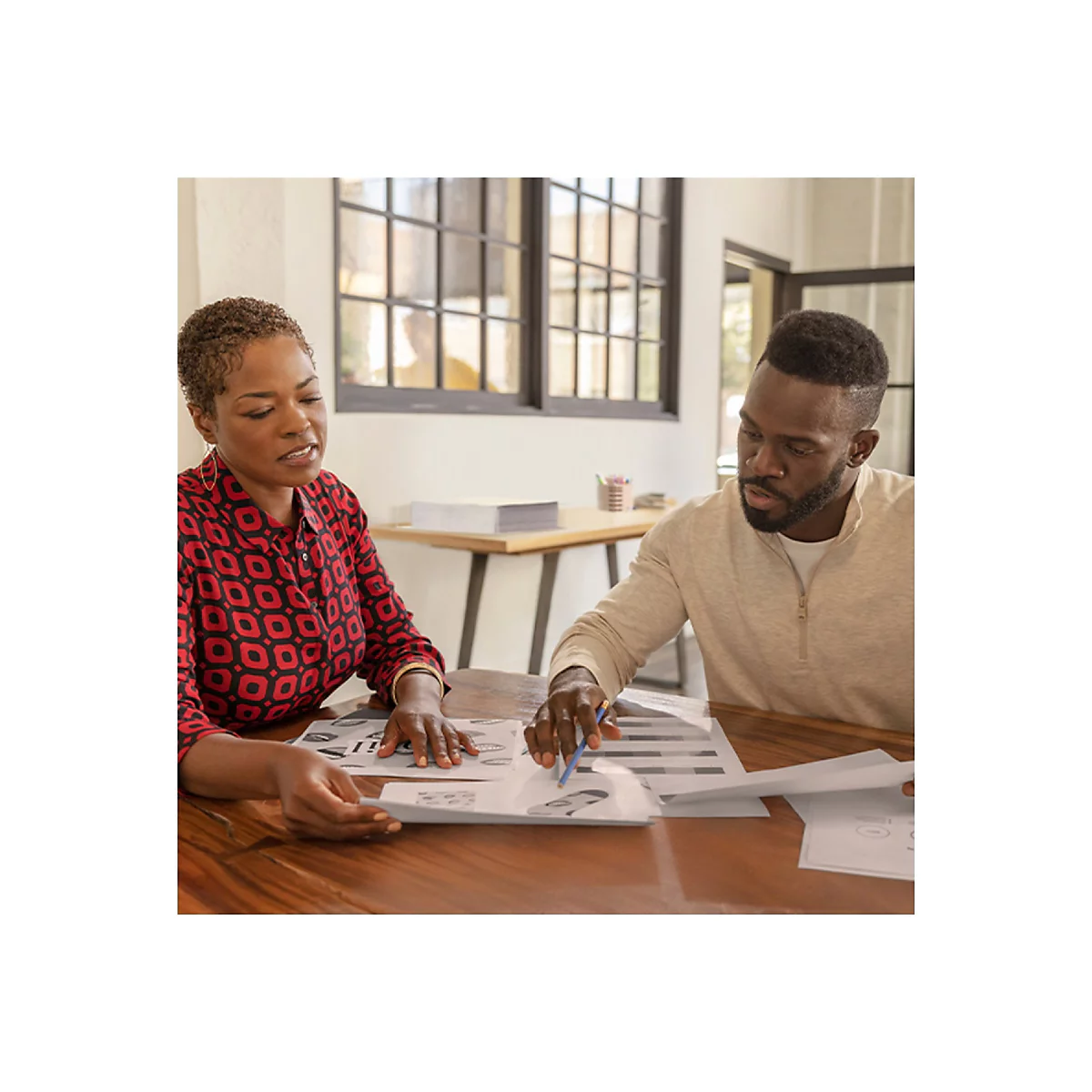 Deux personnes regardent des documents ensemble Ã  une table. Une femme en chemise rouge, un homme en pull beige.