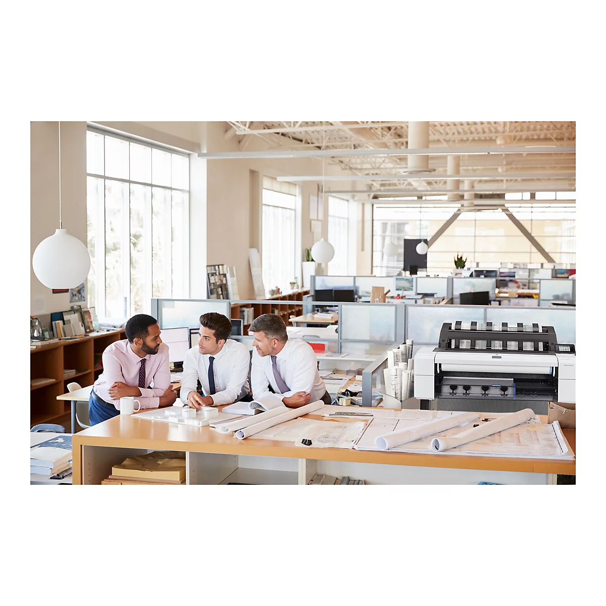 Drei Männer an einem Tisch in einem Büro, die sich über Baupläne beugen und eine große Drucker-Plotter-Maschine betrachten.
