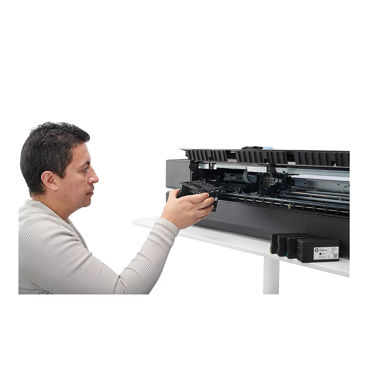 Ein Mann tauscht eine schwarze Tintenpatrone in einem grossen Tintenstrahldrucker aus. Der Drucker und die Tintenpatrone sind auf einem weissen Tisch vor weissem Hintergrund.
