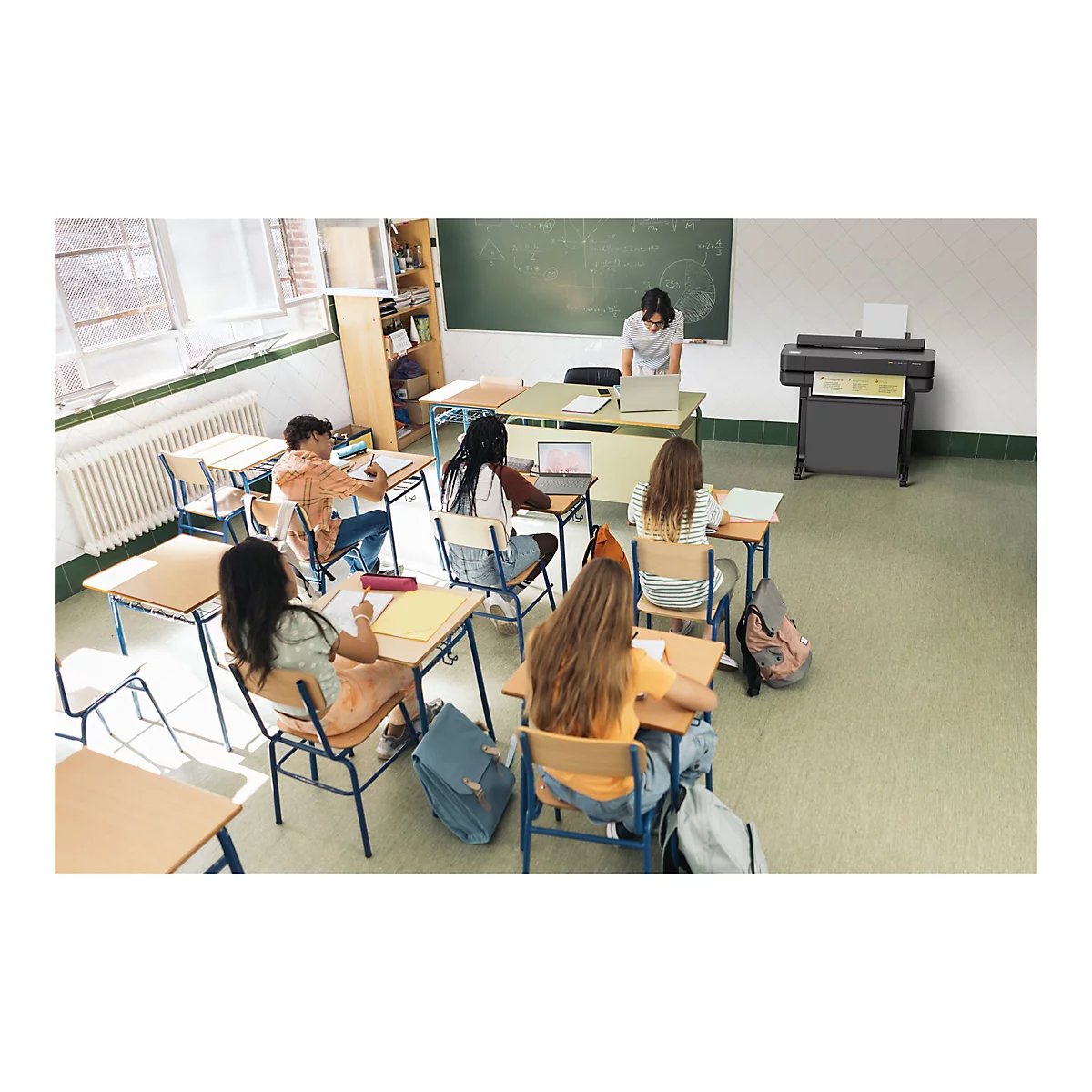 Ein Klassenzimmer, in dem Schüler an ihren Tischen arbeiten. Eine Lehrerin steht am Whiteboard. Rechts steht ein großer Drucker.