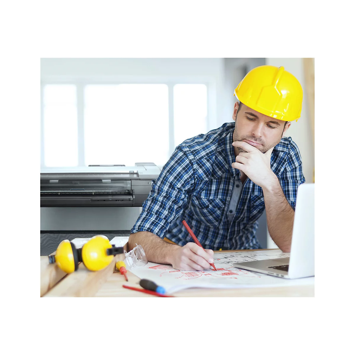 Ein Mann mit gelbem Helm zeichnet auf einem Plan; Laptop, Drucker, Gehörschutz und Brille liegen am Tisch.