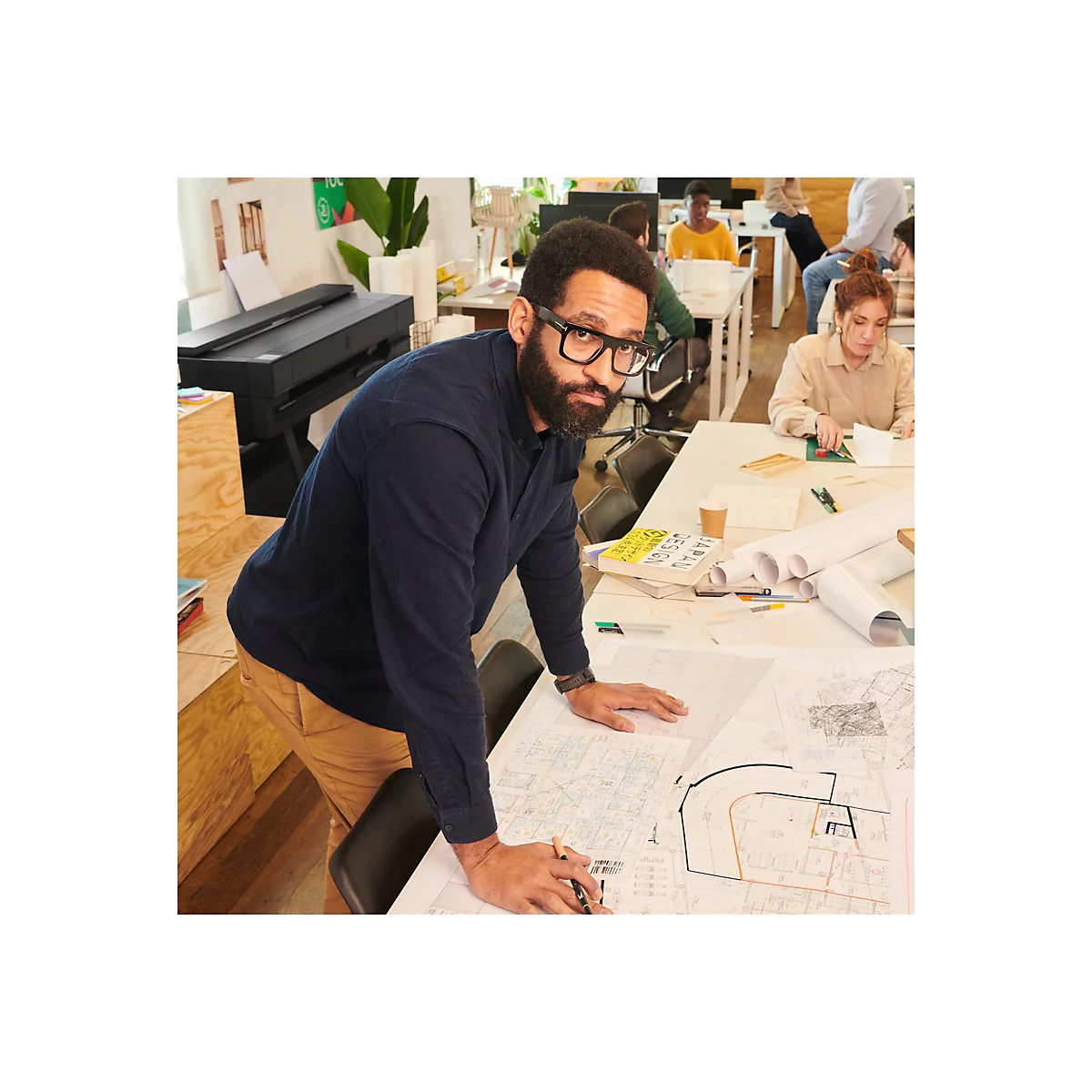 Ein Mann mit Brille beugt sich Ã¼ber BauplÃ¤ne am Tisch, weitere Personen arbeiten im Hintergrund.