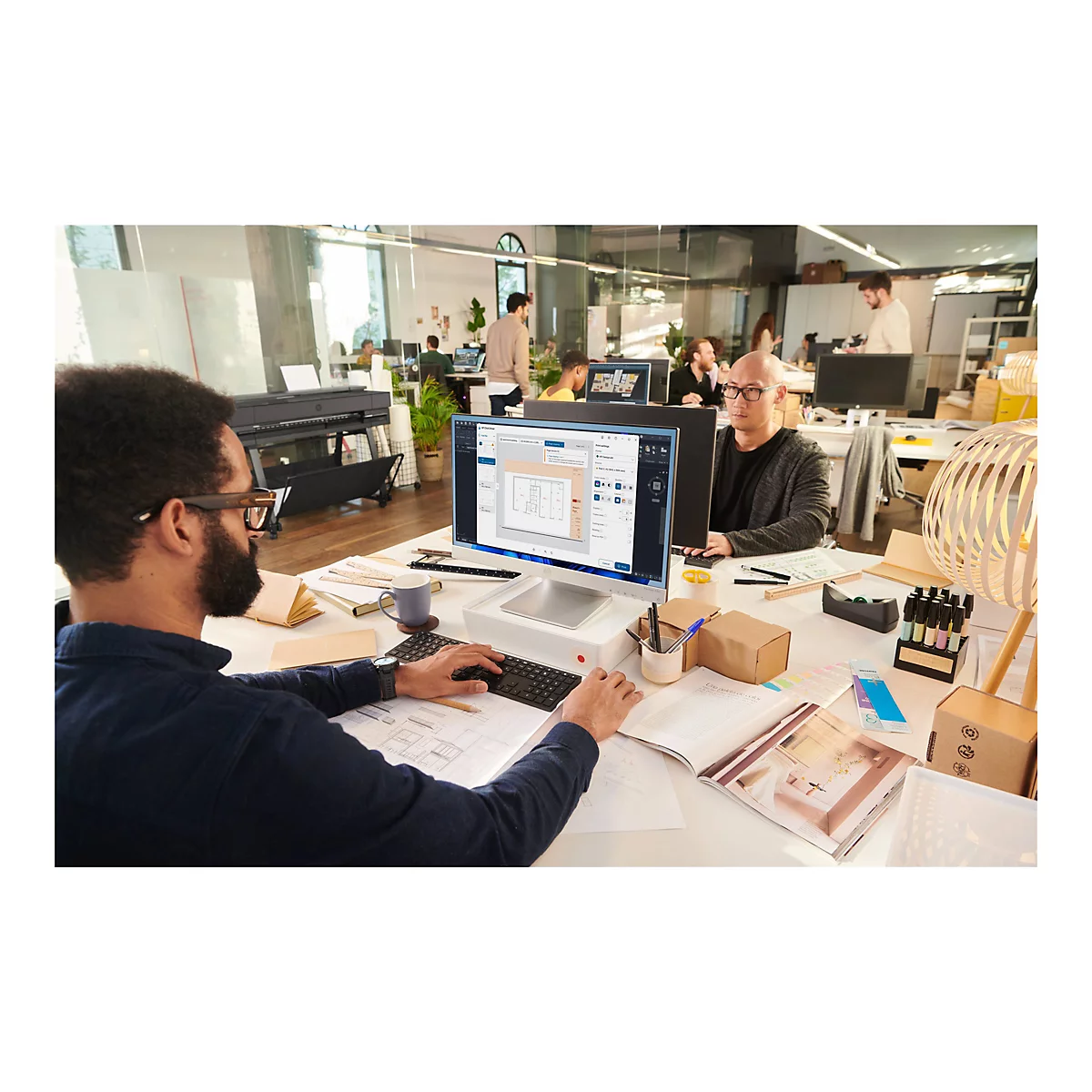 Ein Mann mit Bart sitzt an einem Computer, auf dem ein architektonischer Plan zu sehen ist. Weitere Personen arbeiten im Hintergrund.