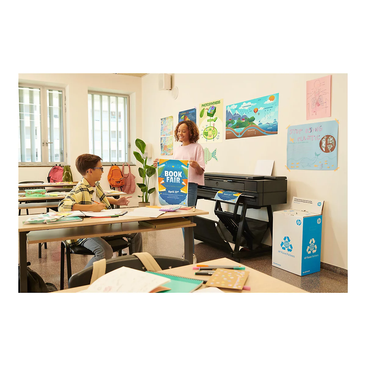 Zwei Schüler in einem Klassenzimmer. Ein Mädchen hält ein Plakat für eine Buchmesse. Auf dem Tisch steht ein Drucker. Auf dem Boden steht eine Recyclingbox.