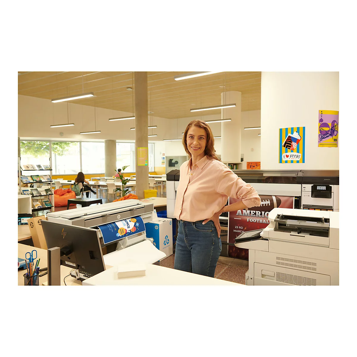 Eine lächelnde Frau steht in einem Büro. Hinter ihr befinden sich Drucker, Plakate und ein Poster. Sie trägt eine rosafarbene Bluse und Jeans.