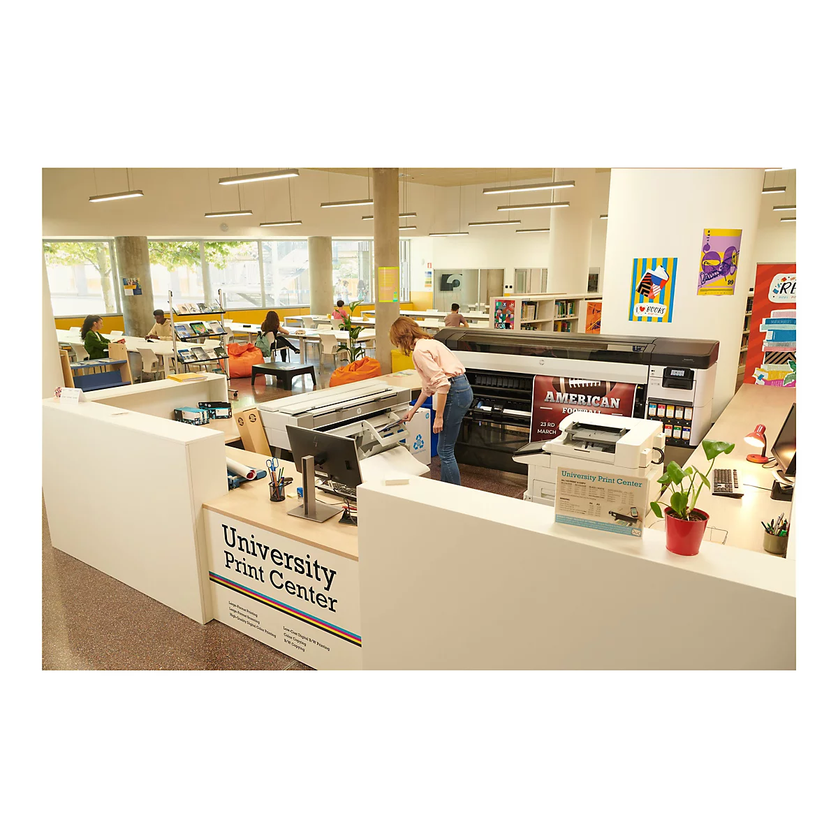 Eine Frau an der Theke des „University Print Center“, befüllt einen Drucker. Im Hintergrund sitzen Leute an Tischen oder in Sitzsäcken.