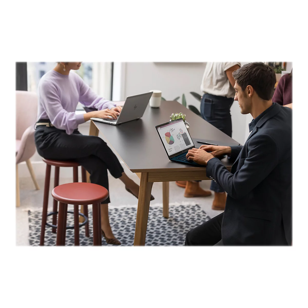 Zwei Personen sitzen an einem Tisch und arbeiten an Laptops. Im Hintergrund stehen weitere Personen.