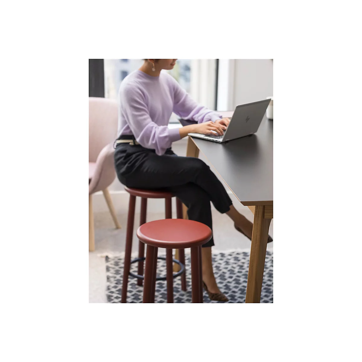 Eine Frau sitzt an einem Schreibtisch und arbeitet an einem Laptop, umgeben von StÃ¼hlen und Teppich.