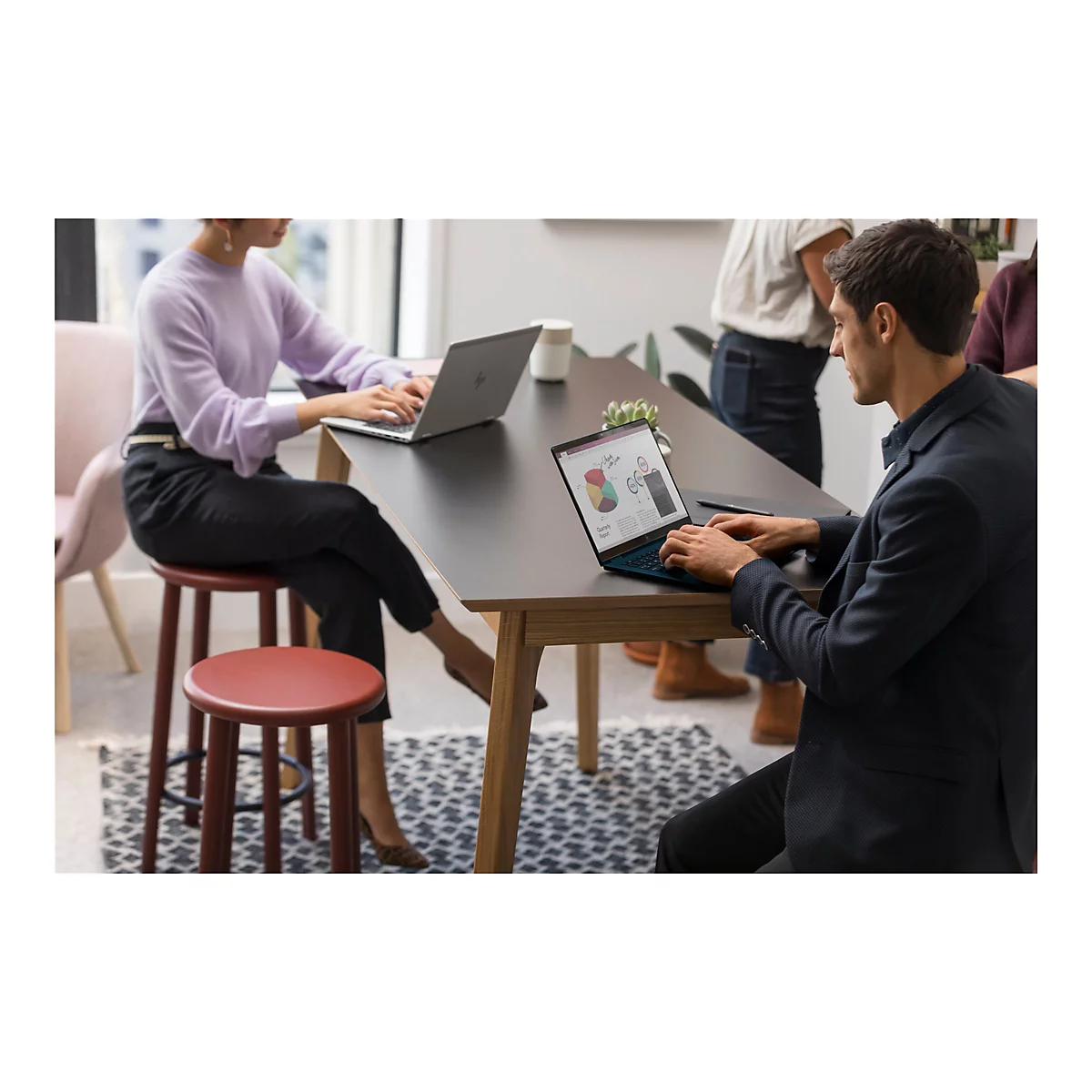 Zwei Personen arbeiten an Laptops an einem Tisch. Ein Mann zeigt Diagramme auf einem Bildschirm. Weitere Personen stehen im Hintergrund.