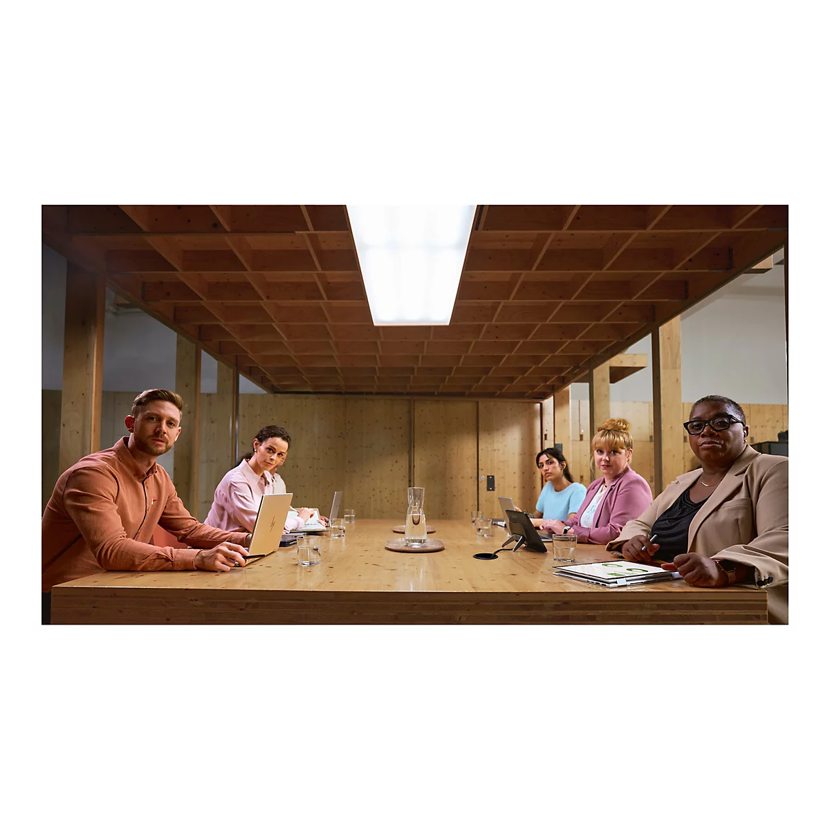 Eine Gruppe von sechs Personen sitzt an einem großen Holztisch in einem Konferenzraum. Sie arbeiten an Laptops und schauen in die Kamera.