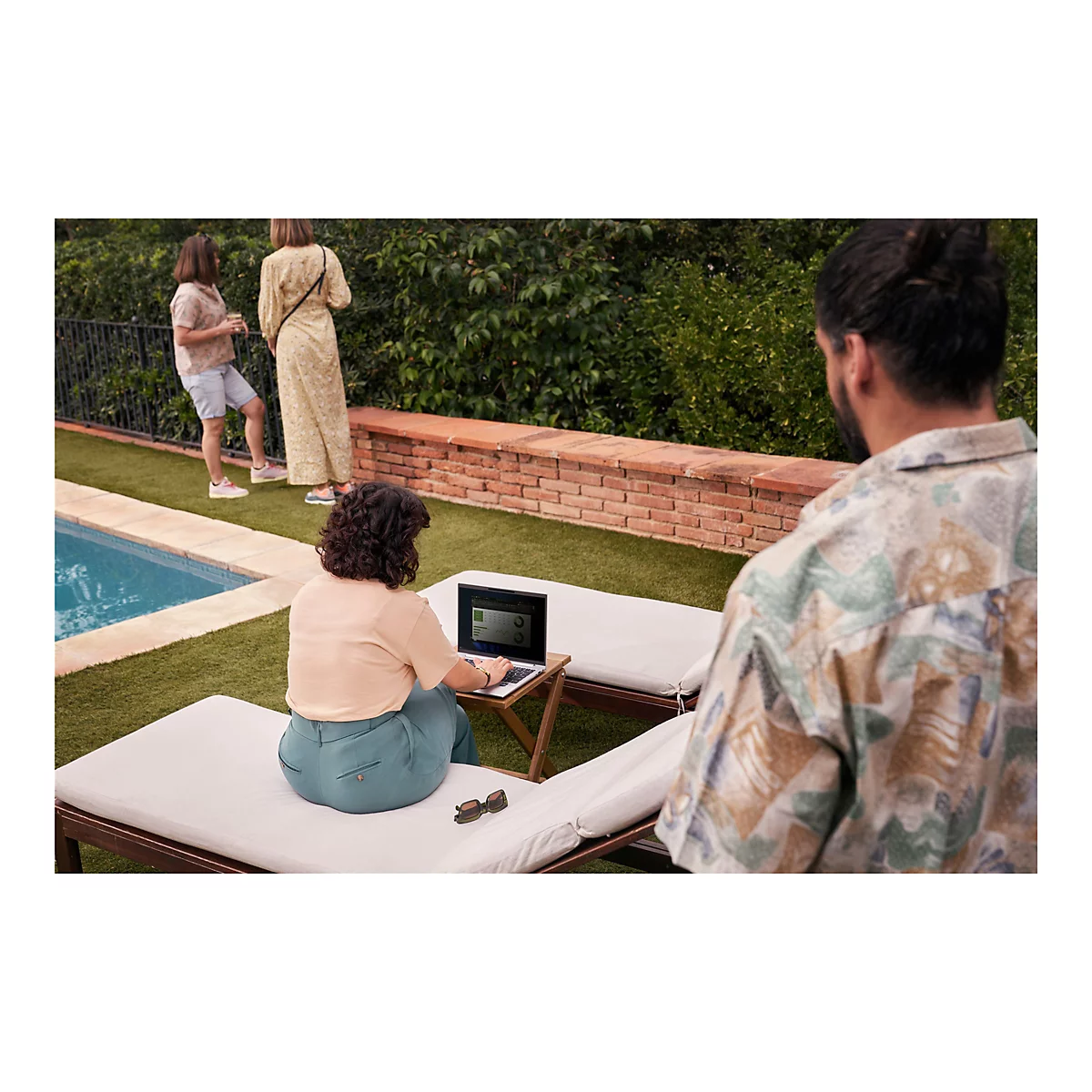 Eine Frau sitzt auf einer Liege und arbeitet mit einem Laptop, im Hintergrund weitere Personen am Pool.