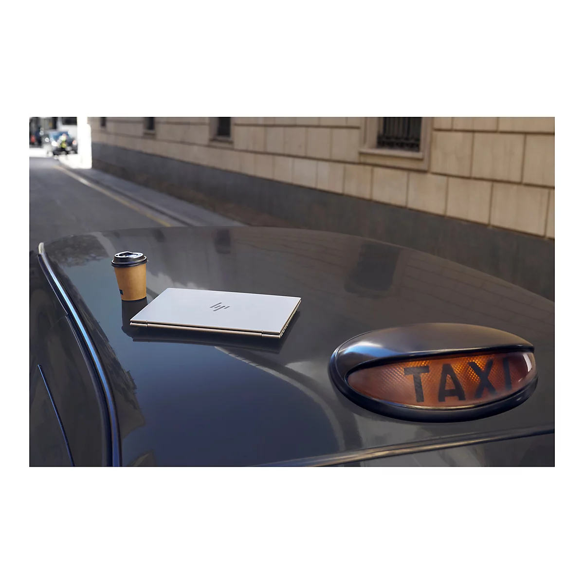 Ein Laptop und ein Pappbecher stehen auf einem Taxi. Im Hintergrund ein Straßenzug.
