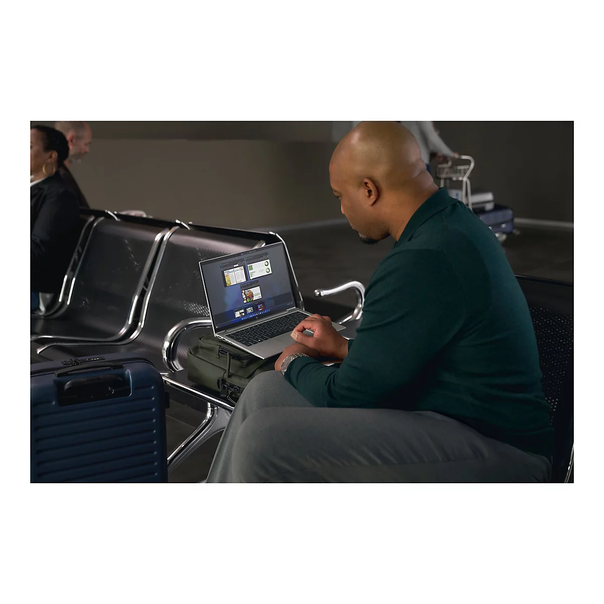 Ein Mann sitzt in einem Wartebereich und arbeitet mit seinem Laptop. Neben ihm ist Gepäck platziert.