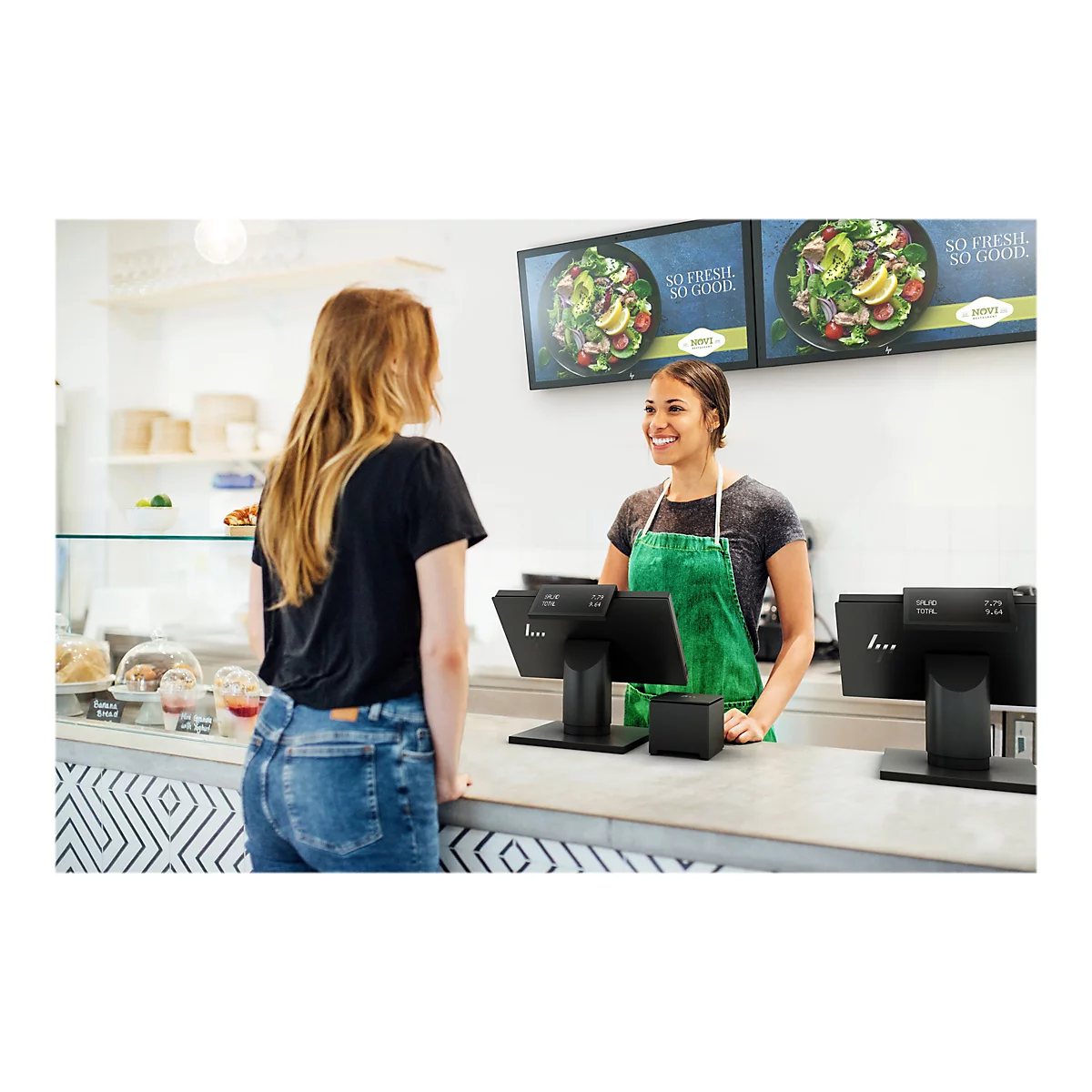 Eine Kundin bestellt an der Theke in einem Restaurant. Die VerkÃ¤uferin lÃ¤chelt und blickt die Kundin an. Hinter ihr sind zwei Bildschirme mit Salatwerbung.