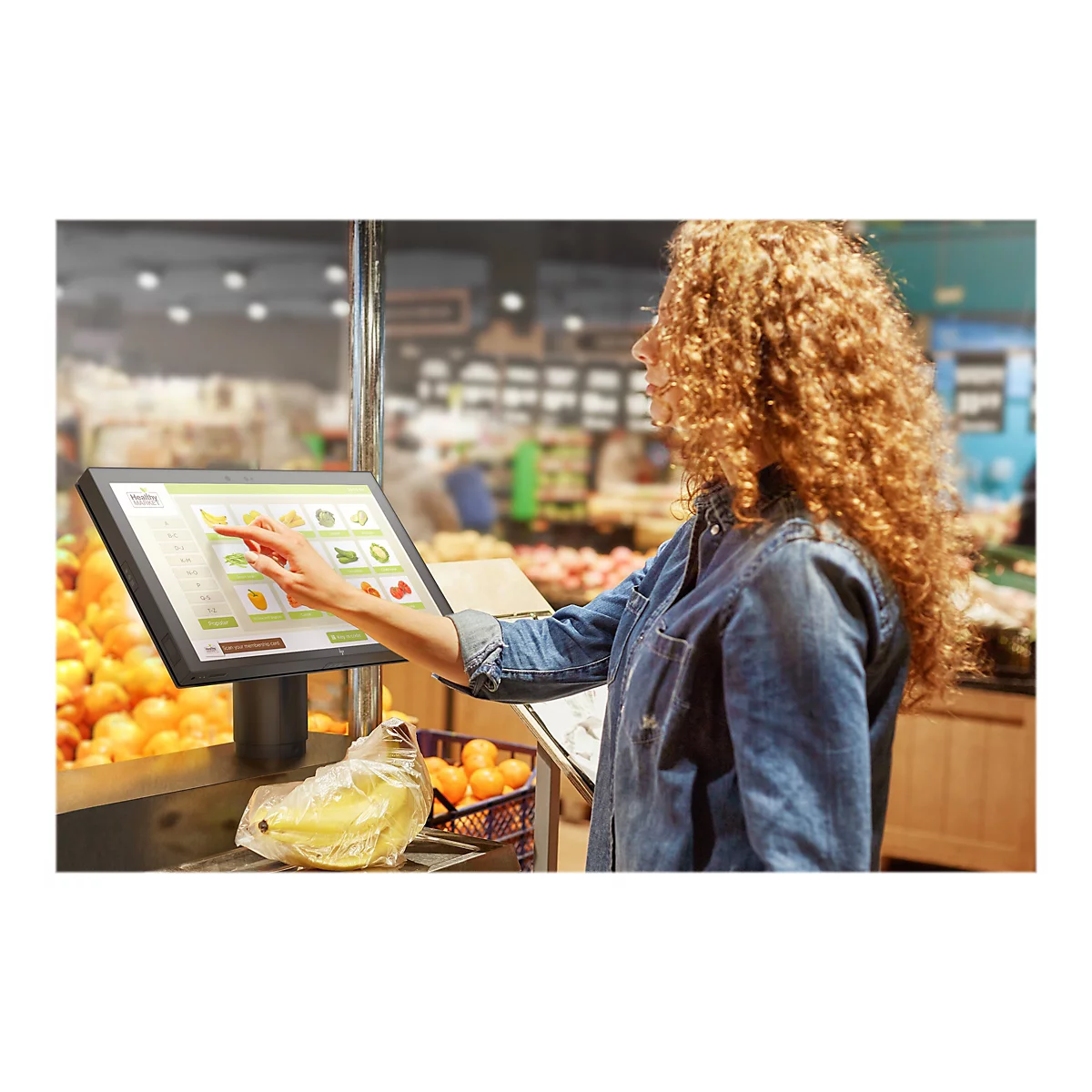 Eine Frau berÃ¼hrt einen Touchscreen in einem Supermarkt. Auf dem Bildschirm werden verschiedene Obst- und GemÃ¼searten angezeigt.