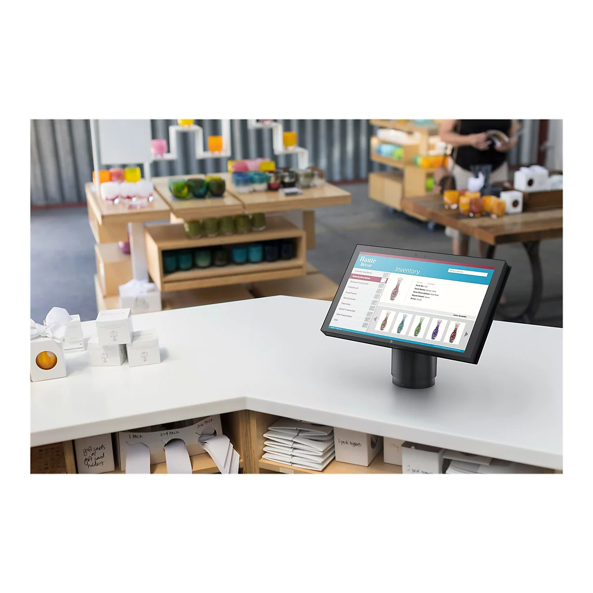 Ein POS-Terminal steht auf einer Theke in einem GeschÃ¤ft. Der Bildschirm zeigt eine Produktliste. Im Hintergrund sind weitere Waren und eine Person zu sehen.
