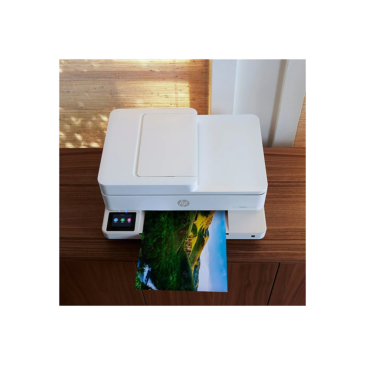 Weißer HP Envy Drucker, druckt ein Farbfoto eines grünen Hügels mit blauem Himmel. Drucker steht auf dunklem Holzschrank.