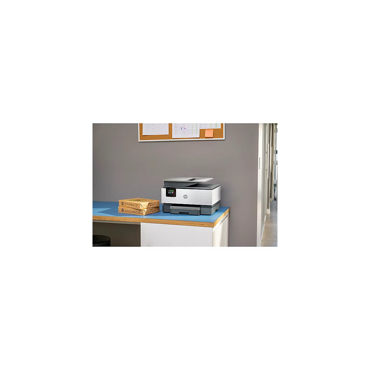 Une imprimante HP grise est posée sur une table bleue, avec 2 paquets de papier à côté. En arrière-plan, un tableau d'affichage.