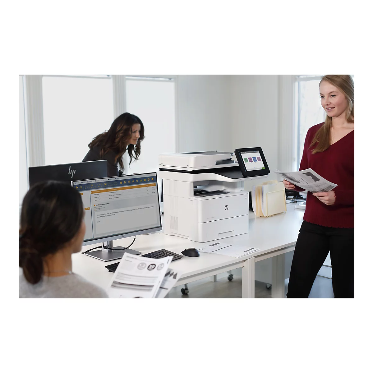 Büro mit drei Personen. Ein Drucker steht auf einem Schreibtisch, eine Frau hält Papier, eine andere schaut auf einen Monitor.