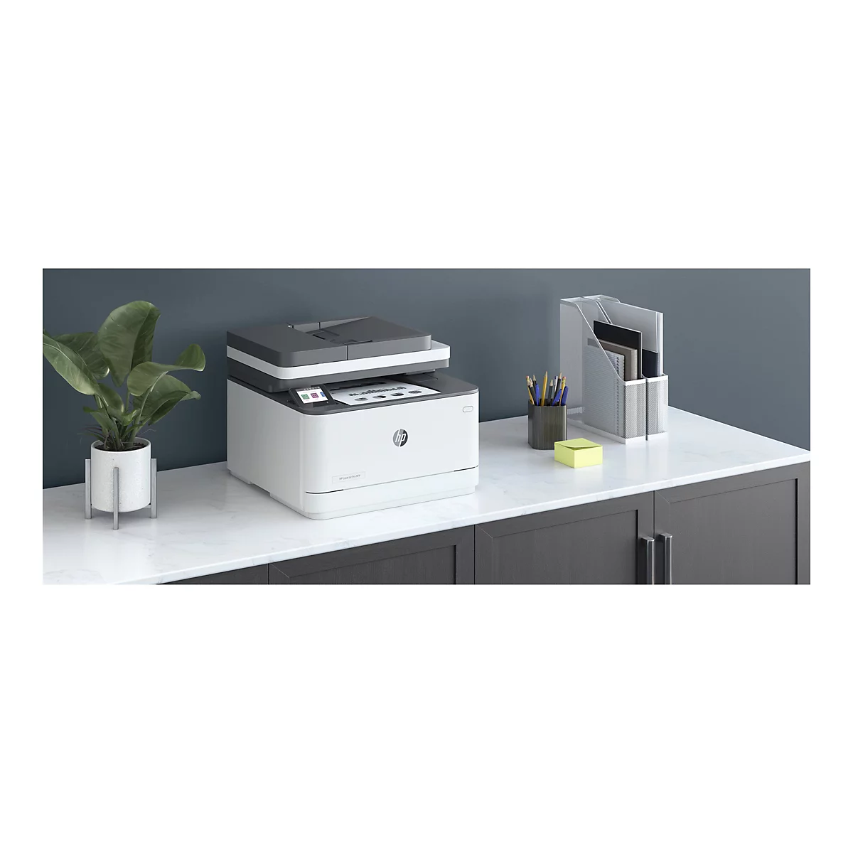 Ein weiß-grauer HP-Laserdrucker steht auf einer weißen Oberfläche. Im Hintergrund sind Büromaterialien und eine Topfpflanze zu sehen.
