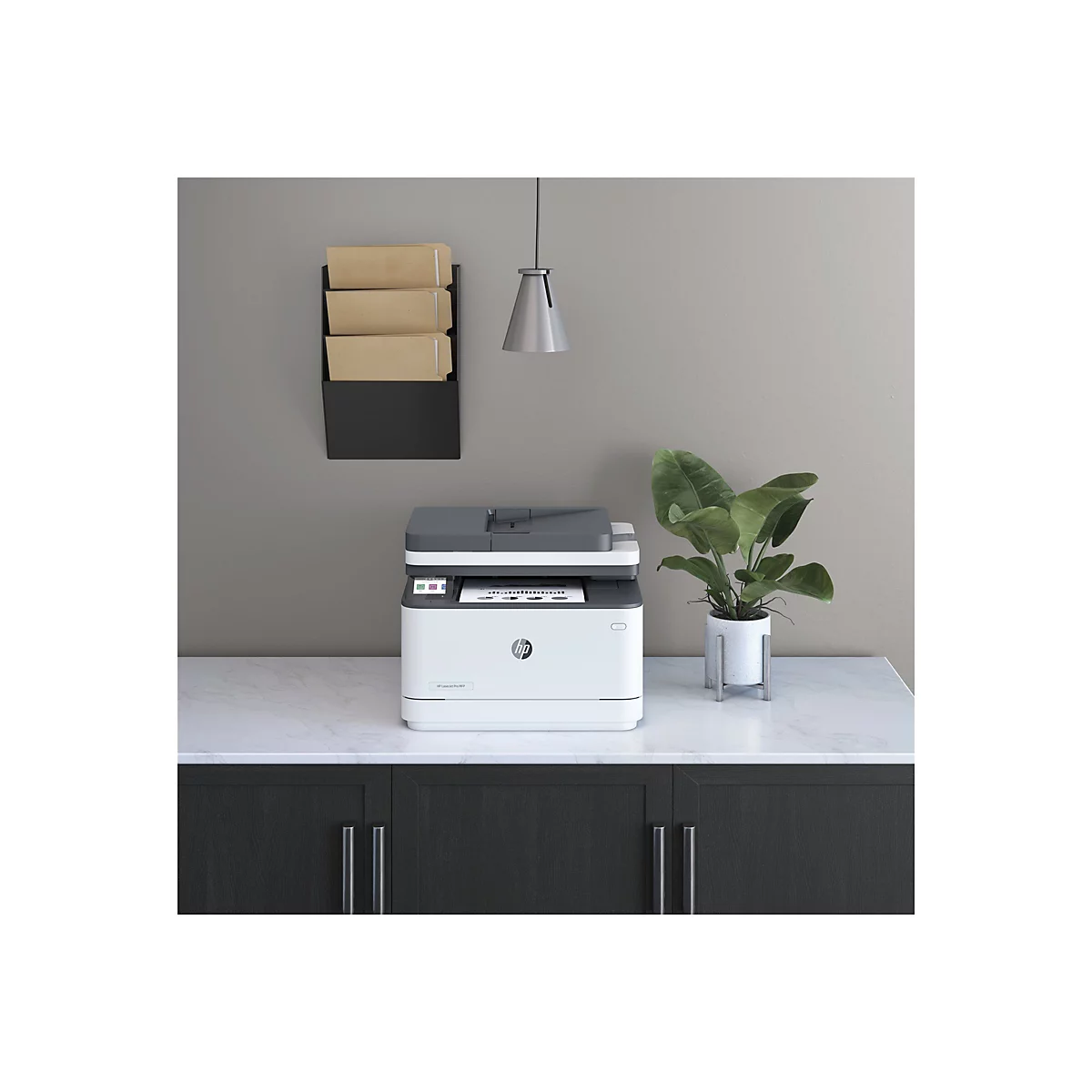 Ein weißer HP-Laserdrucker steht auf einer hellen Oberfläche. Rechts daneben steht eine Topfpflanze, an der Wand ein schwarzes Ablagesystem.