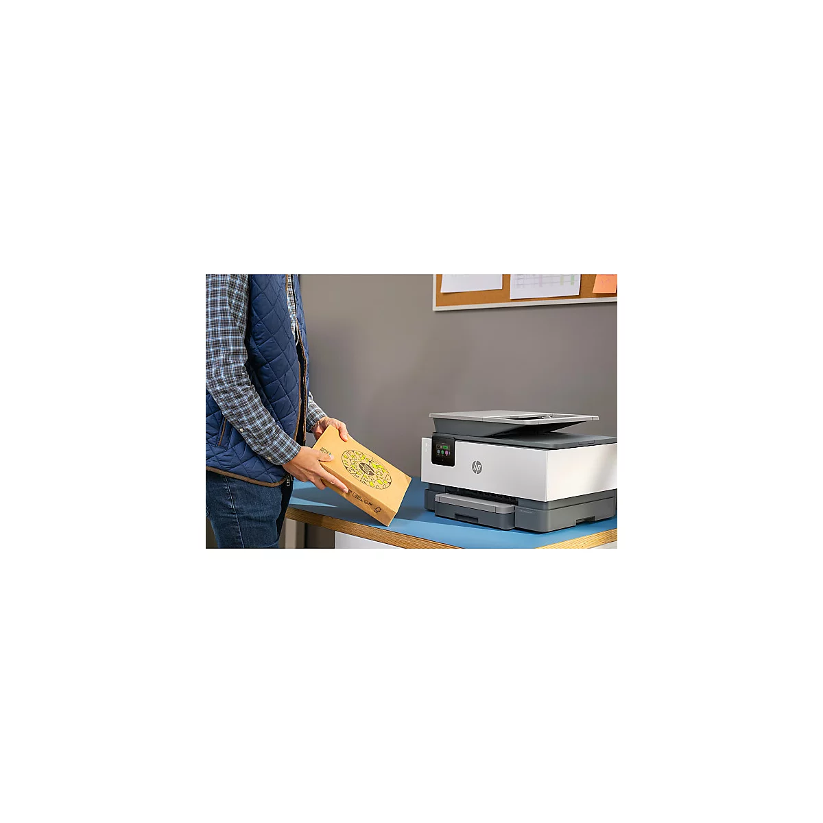 Een persoon plaatst een printercartridge op een tafel naast een HP multifunctionele printer. Op de tafel ligt een bruine doos.