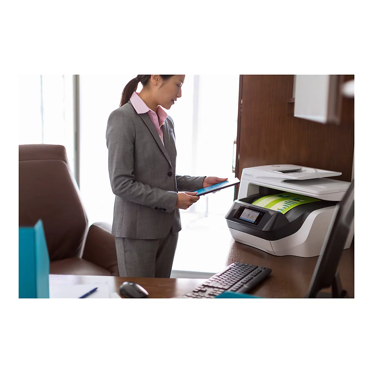 Eine Frau in einem grauen Anzug steht an einem Schreibtisch und hält ein Tablet vor einem Drucker. Der Drucker, eine HP OfficeJet Pro 7740, ist geöffnet und druckt ein Dokument.