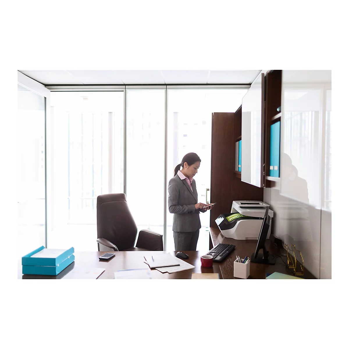Eine Geschäftsfrau in einem Anzug, die in ihrem Büro auf ein Smartphone schaut. Sie steht an ihrem Schreibtisch in einem hellen Büro mit Fenstern.