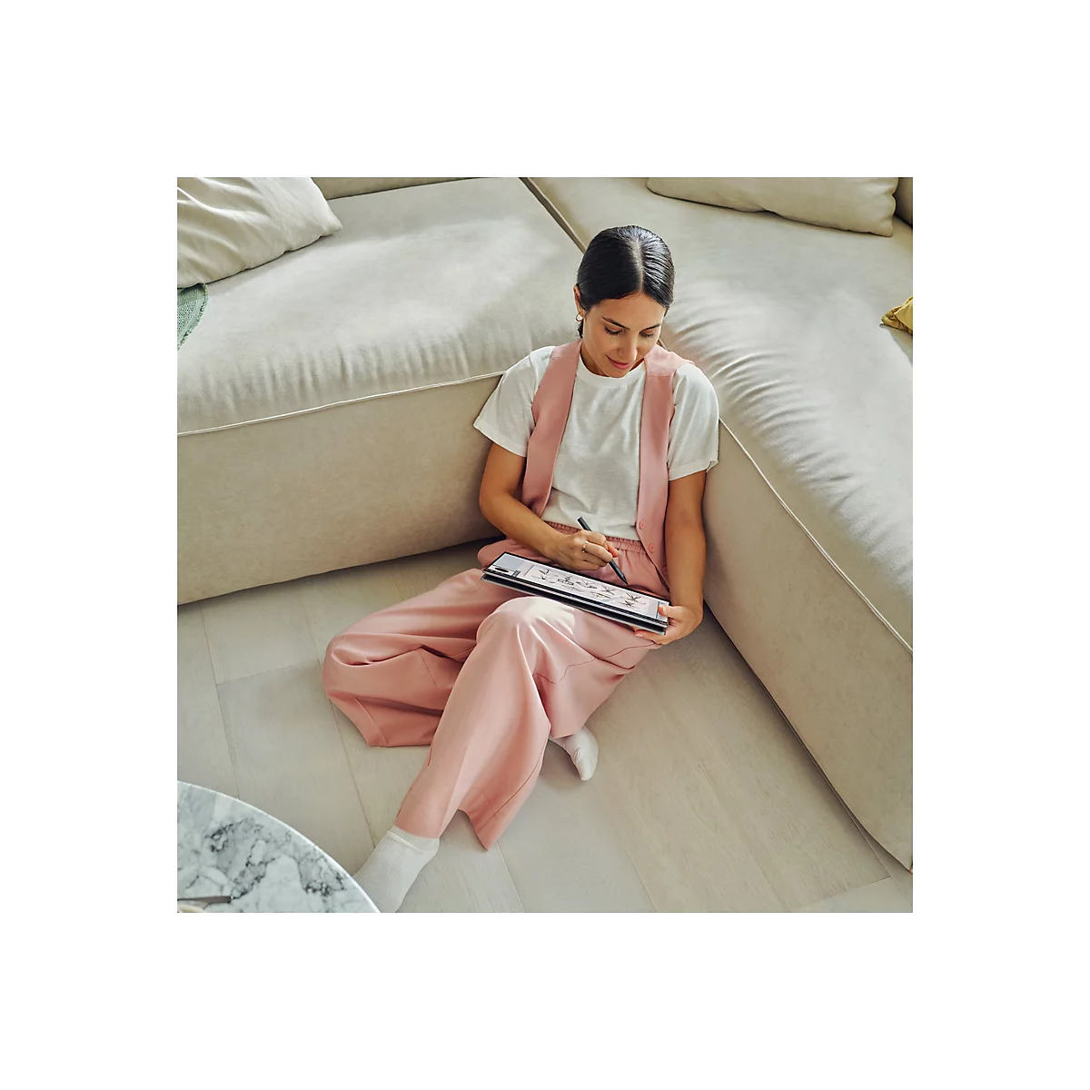 Eine Frau in rosa Hose und Weste sitzt auf dem Boden und zeichnet mit einem Stift auf einem Tablet.