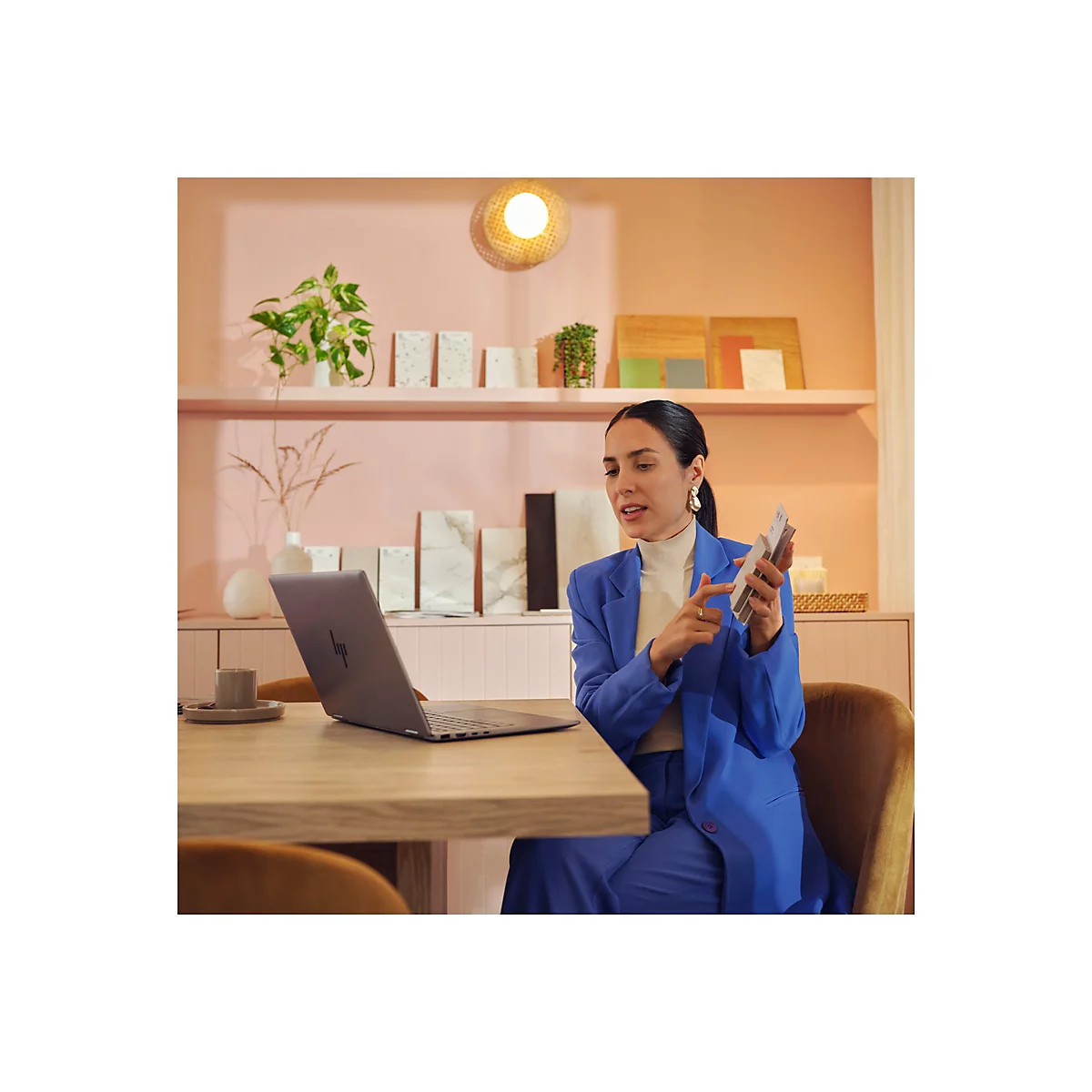 Eine Frau im blauen Anzug sitzt an einem Tisch und präsentiert Farbmuster. Auf dem Tisch befinden sich ein Laptop und eine Kaffeetasse.