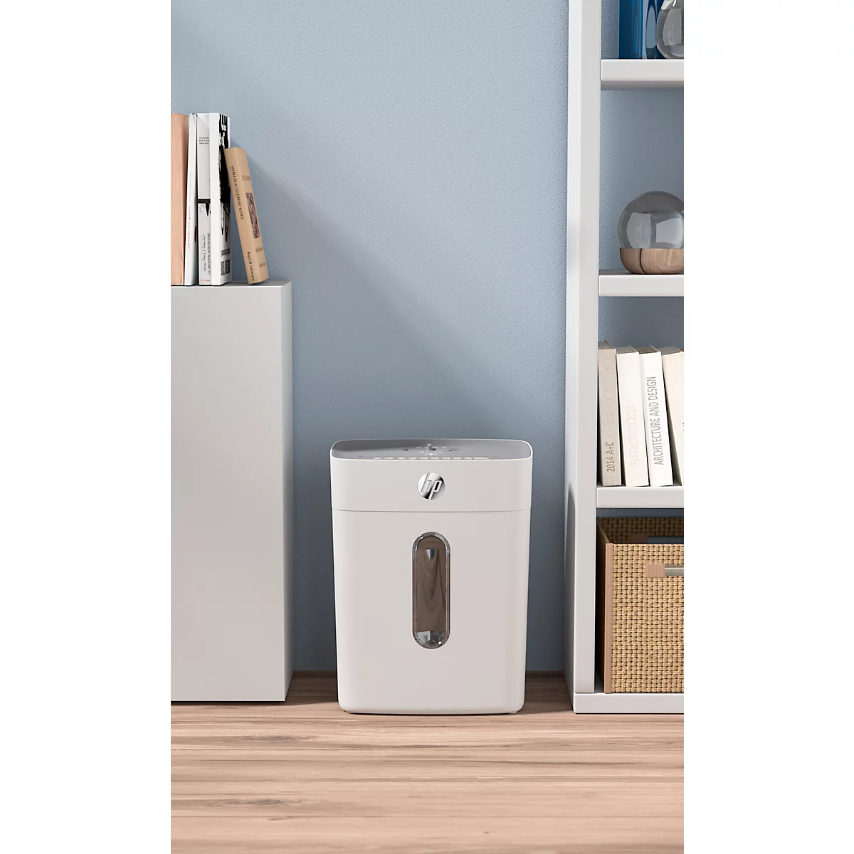 White paper shredder in front of a light blue background. A shelf on the right, a cabinet on the left.