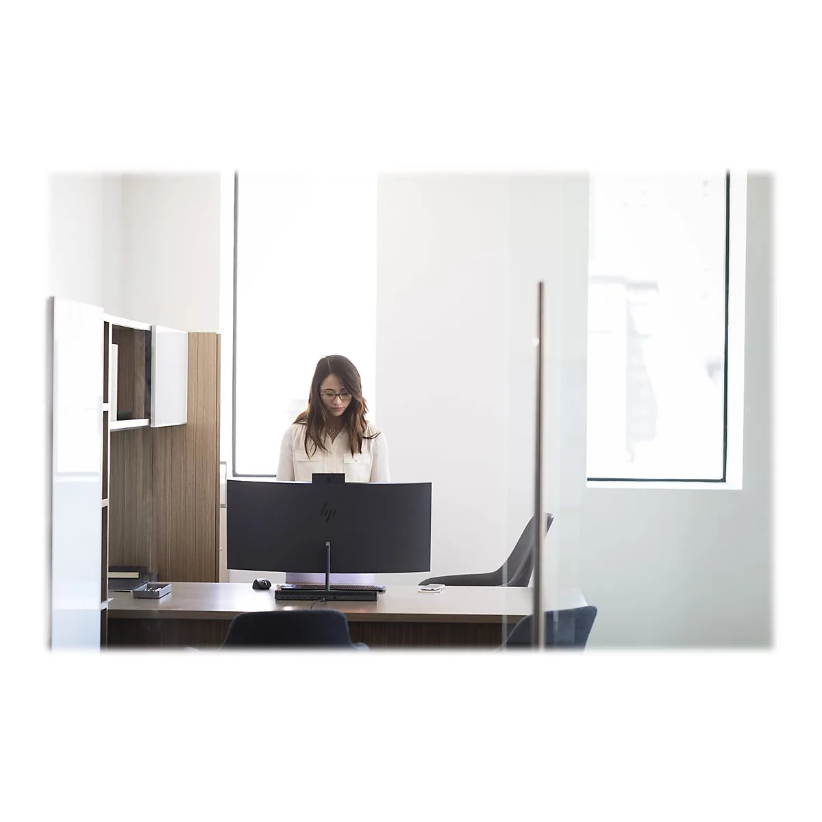 Eine Frau mit Brille sitzt an einem Schreibtisch und arbeitet an einem groÃŸen Computerbildschirm in einem modernen BÃ¼ro.