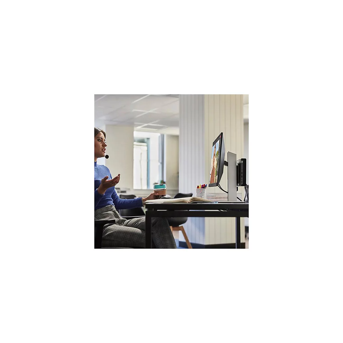 Eine Frau mit Headset sitzt vor einem Monitor, wÃ¤hrend sie einen Becher hÃ¤lt. Auf dem Monitor ist ein Videoanruf zu sehen.
