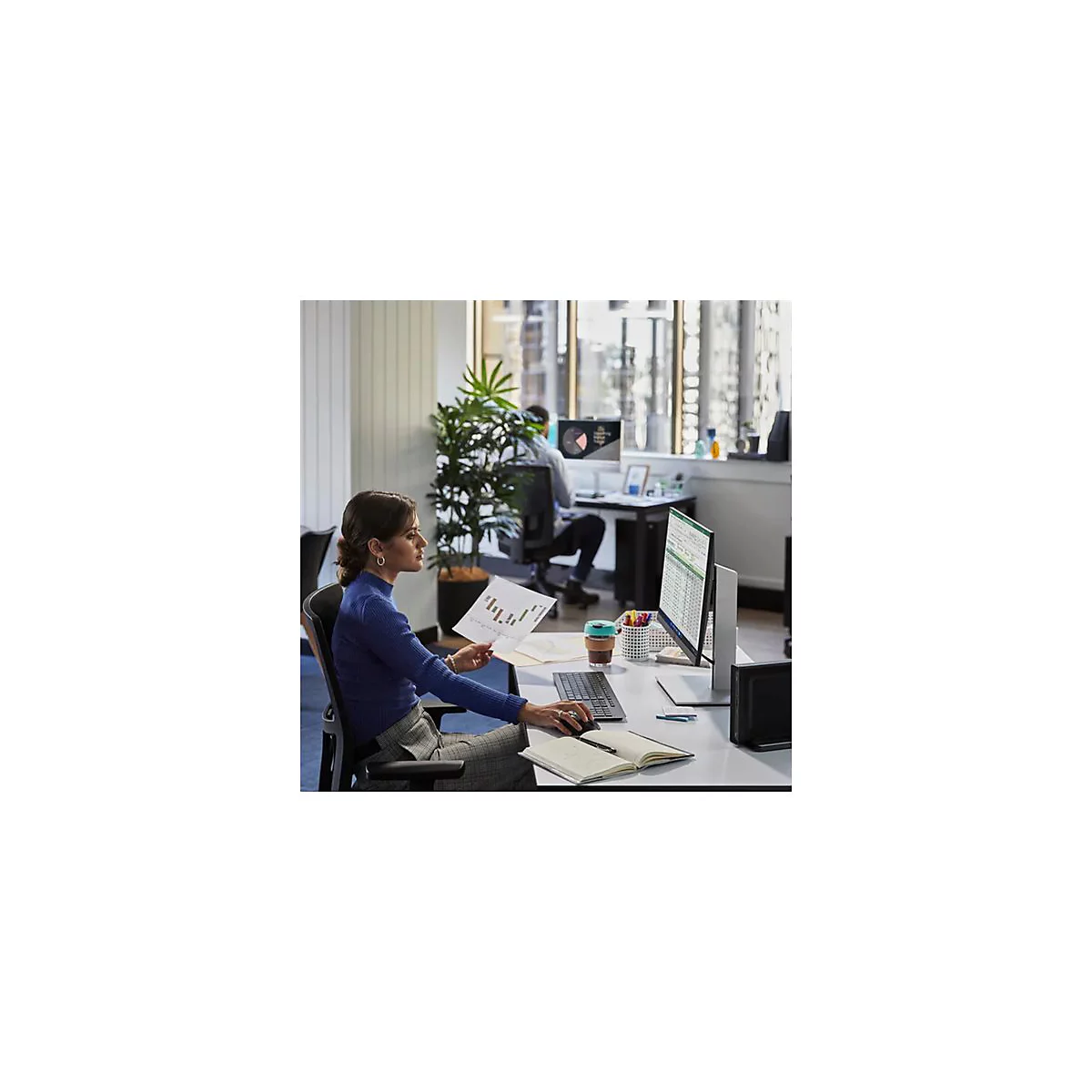 Eine Frau sitzt an einem Schreibtisch und betrachtet Diagramme. Auf dem Tisch befindet sich ein Computerbildschirm und eine Tastatur.
