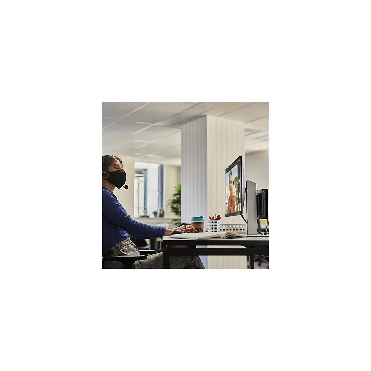 Eine Frau mit Maske und Headset sitzt am Computer, sie nimmt an einer Videokonferenz teil.