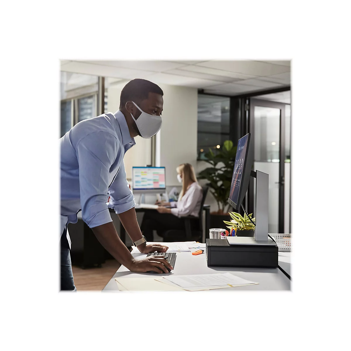 Ein Mann mit Maske arbeitet an einem Computer im BÃ¼ro. Eine weitere Person sitzt im Hintergrund.