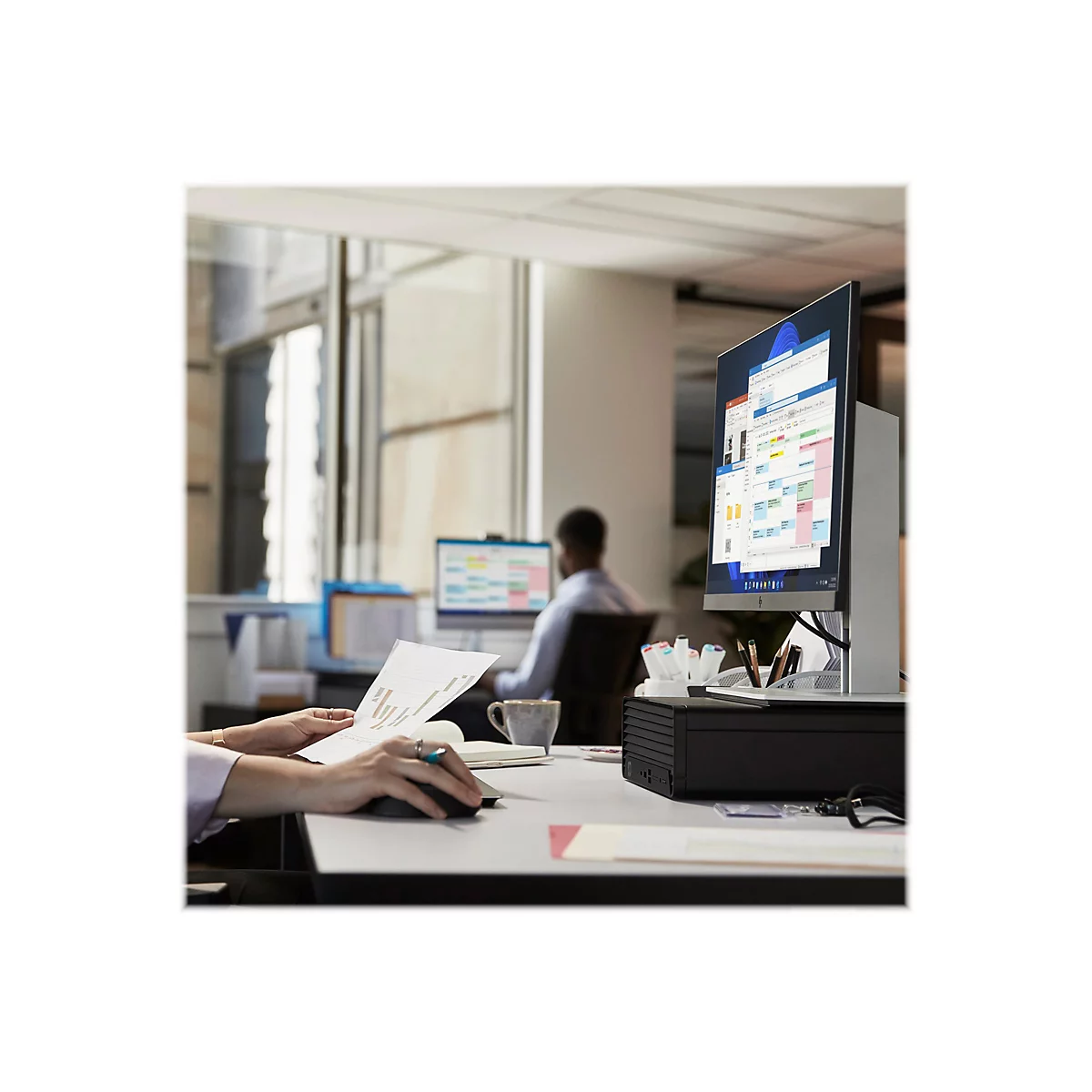 Person bearbeitet Dokumente am Computer im BÃ¼ro. Im Hintergrund weitere Personen am Arbeitsplatz.