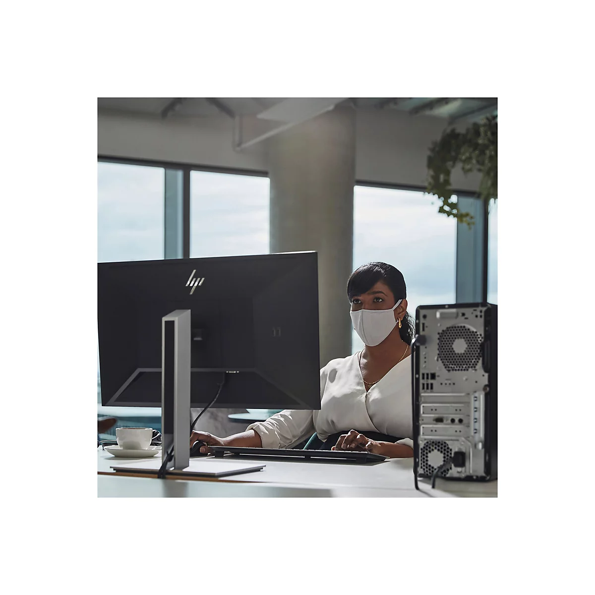 Eine Frau mit Maske sitzt an einem Computer mit groÃŸem Bildschirm, einem PC und einer Tasse Kaffee.