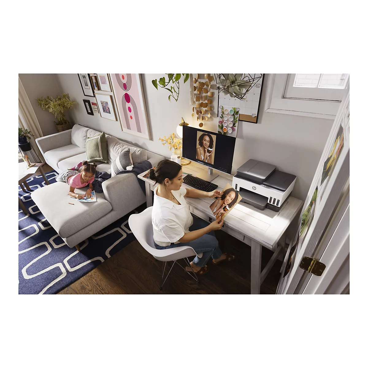 Une femme est assise à un bureau, regardant une photo d'elle-même sur un écran d'ordinateur. Un enfant dessine sur un canapé.