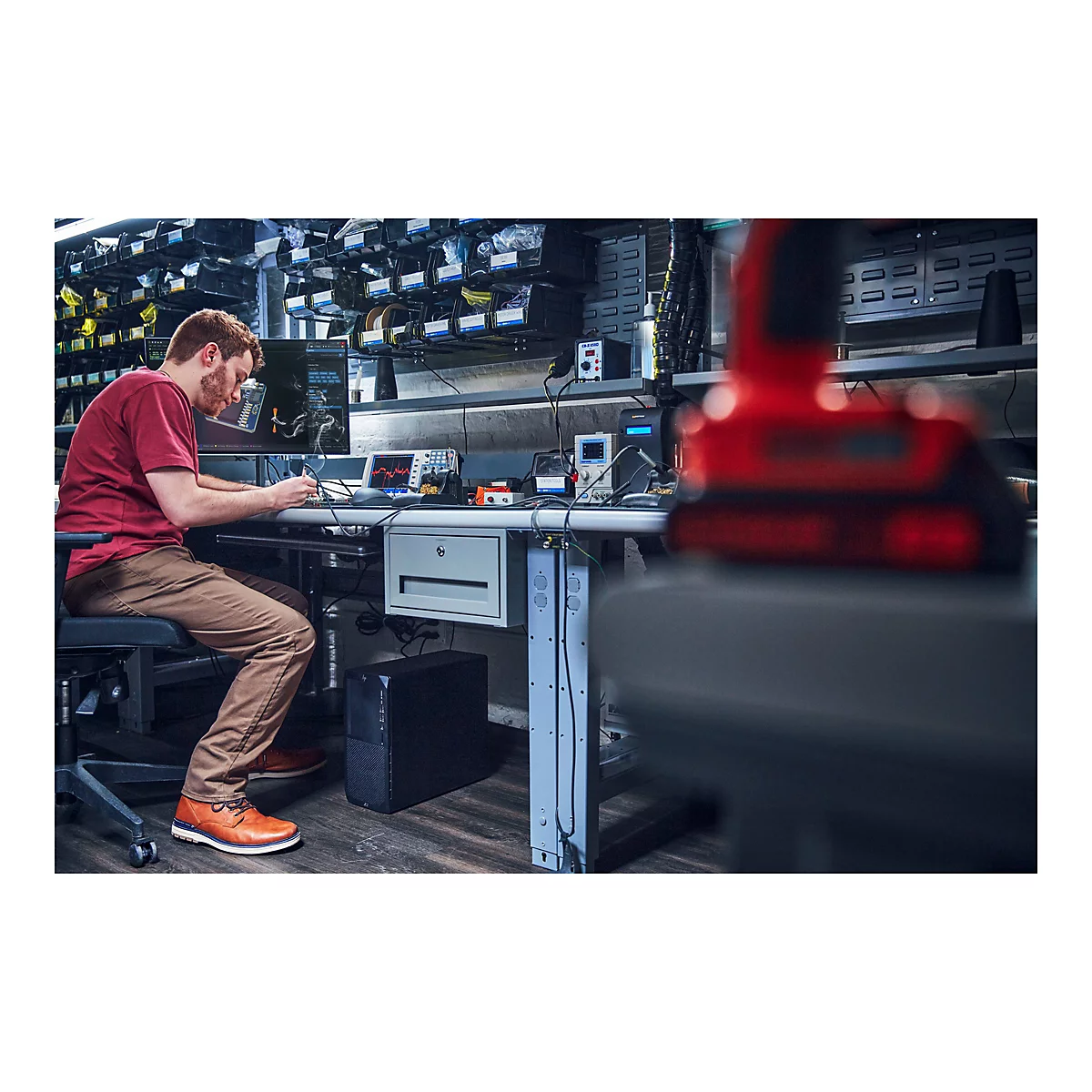 Ein Mann arbeitet an einem Schreibtisch mit elektronischen GerÃ¤ten, wÃ¤hrend im Vordergrund ein rotes Werkzeug unscharf ist.