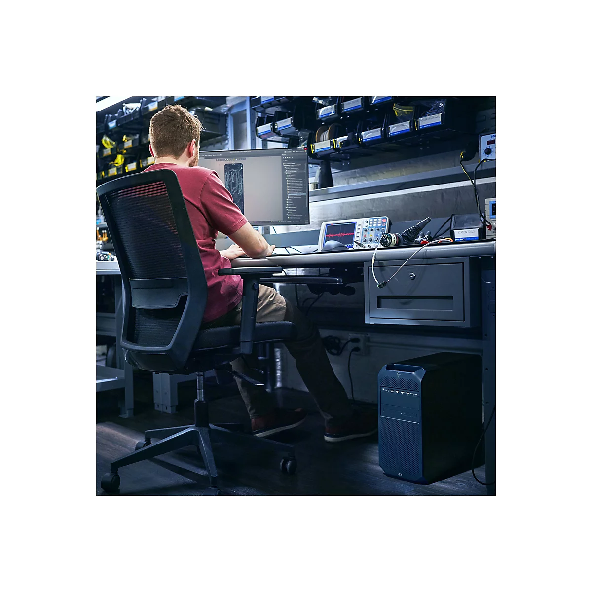 Ein Mann sitzt an einem Schreibtisch und arbeitet am Computer. Er trÃ¤gt ein rotes T-Shirt, der Stuhl ist schwarz.