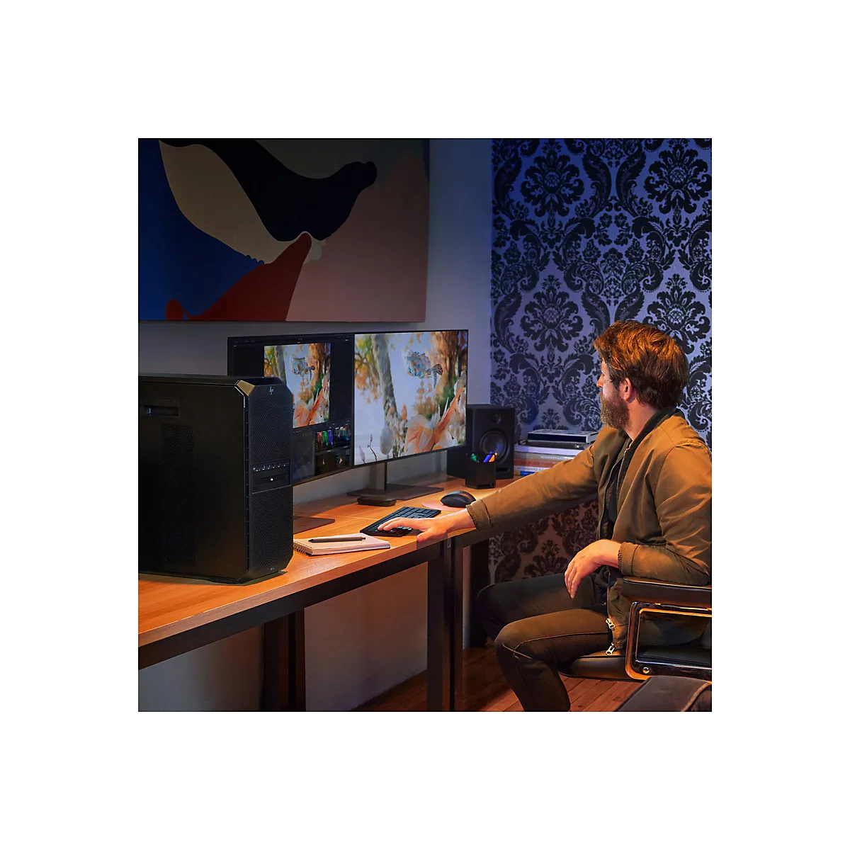 Ein Mann sitzt an einem Schreibtisch und bearbeitet ein Bild auf einem Monitor. Auf dem Schreibtisch stehen Computer, Lautsprecher, Maus und Stift.